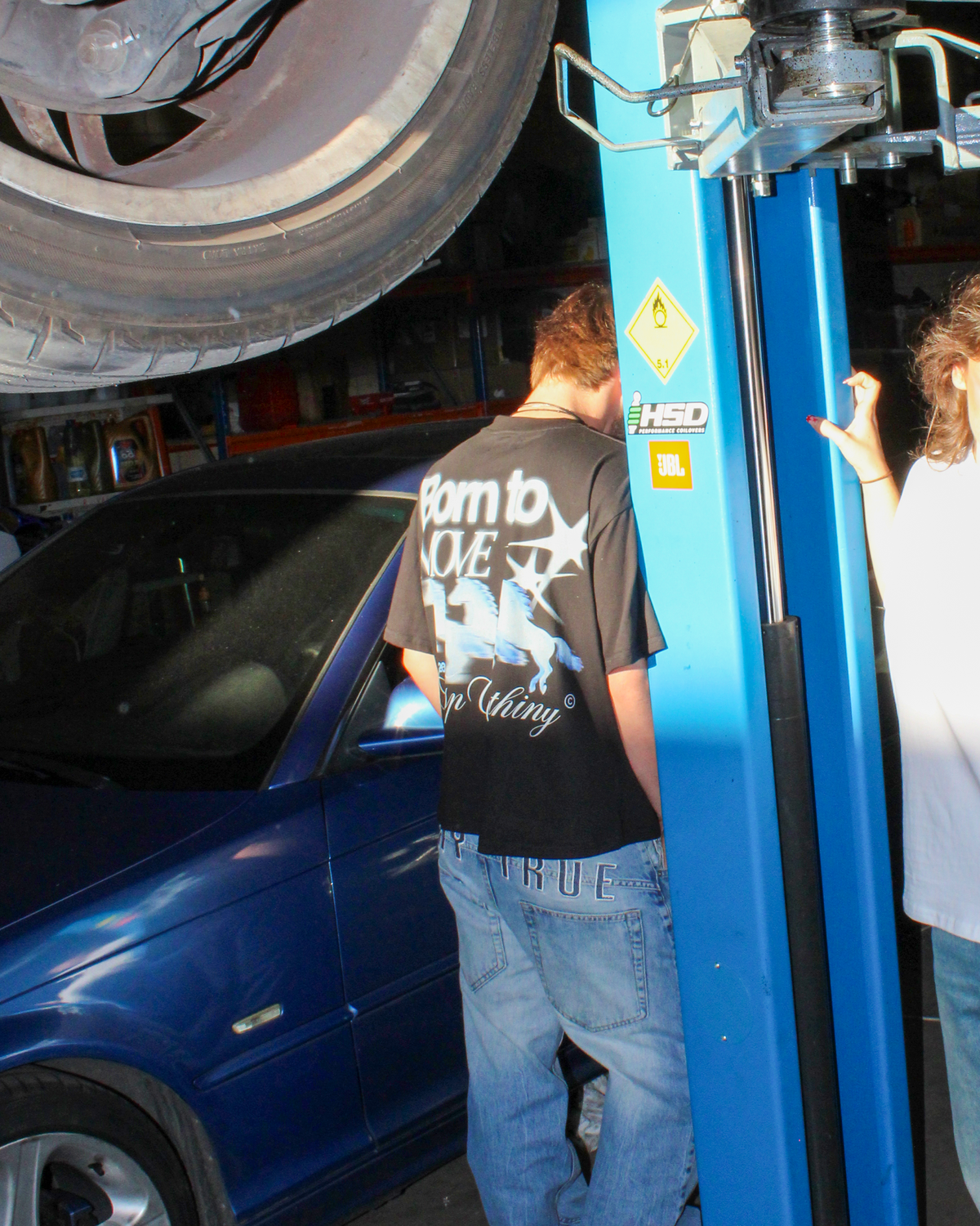Modelo con camiseta oversize negra LIMITLESS de TASP Clothing bajo un elevador de coches, look streetwear